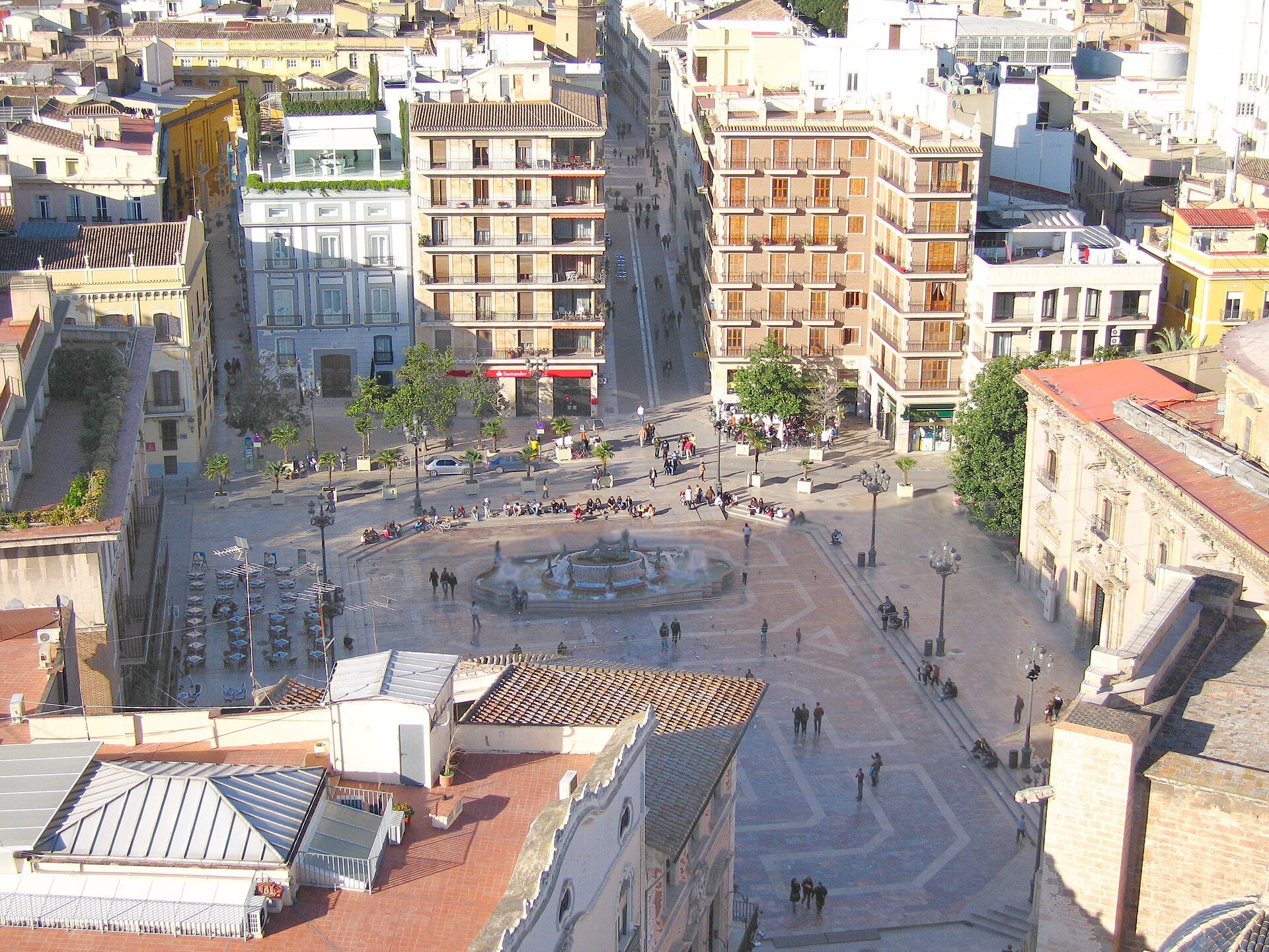 The square seen from above, with its round fountain, café tables and people walking across.