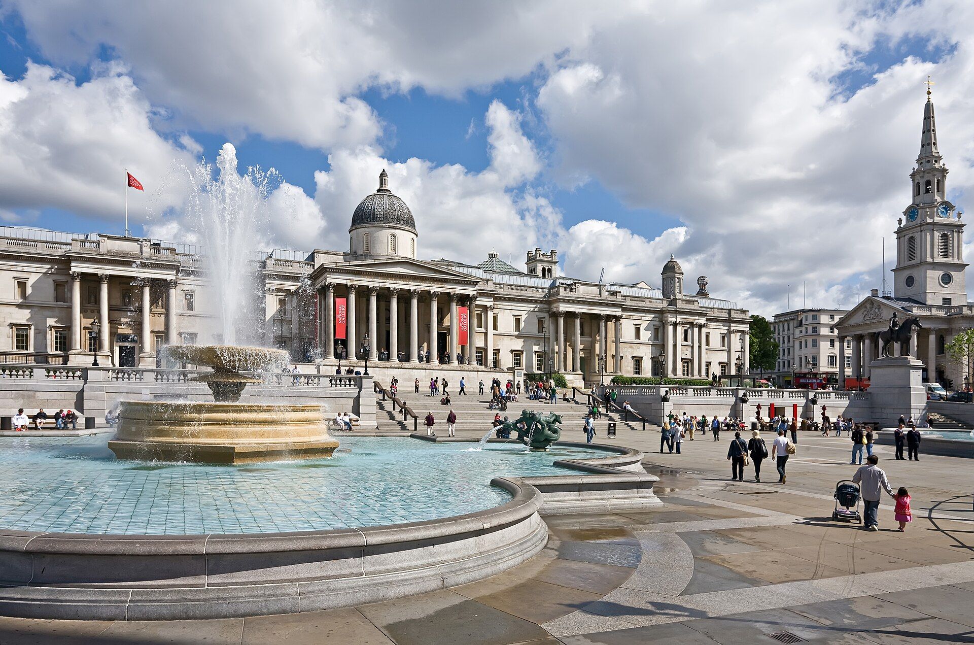 A fountain on Trafalgar Square in sunshine, with the National Gallery and St Martin-in-the-Fields behind.
